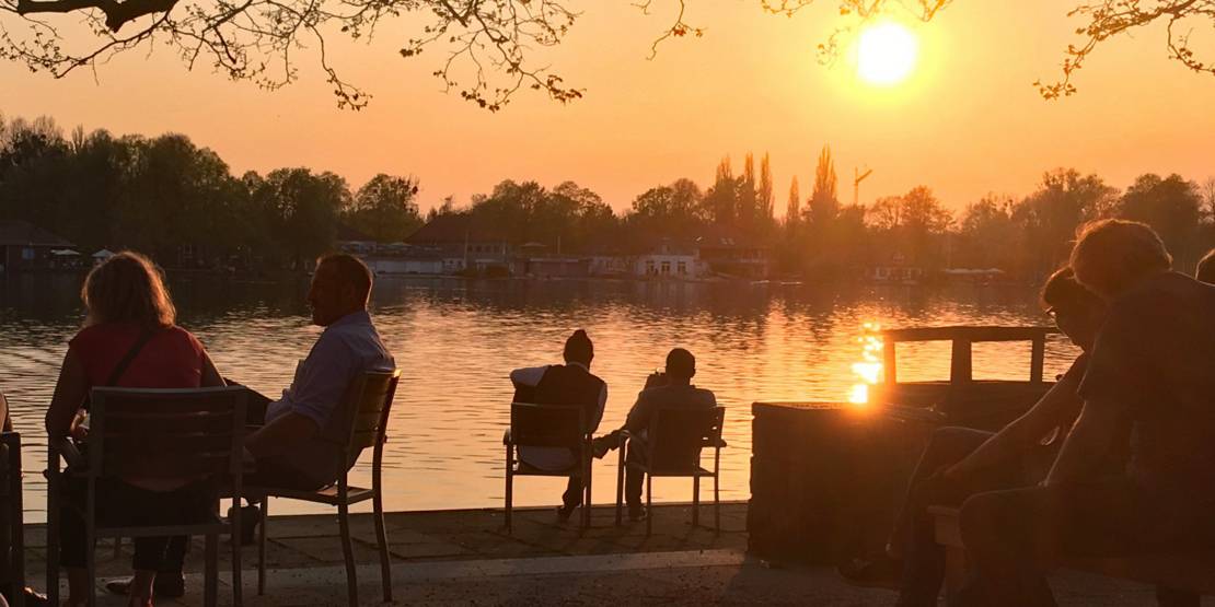 Menschen sitzen auf Stühlen an einem Seeufer und betrachten die untergehende Sonne